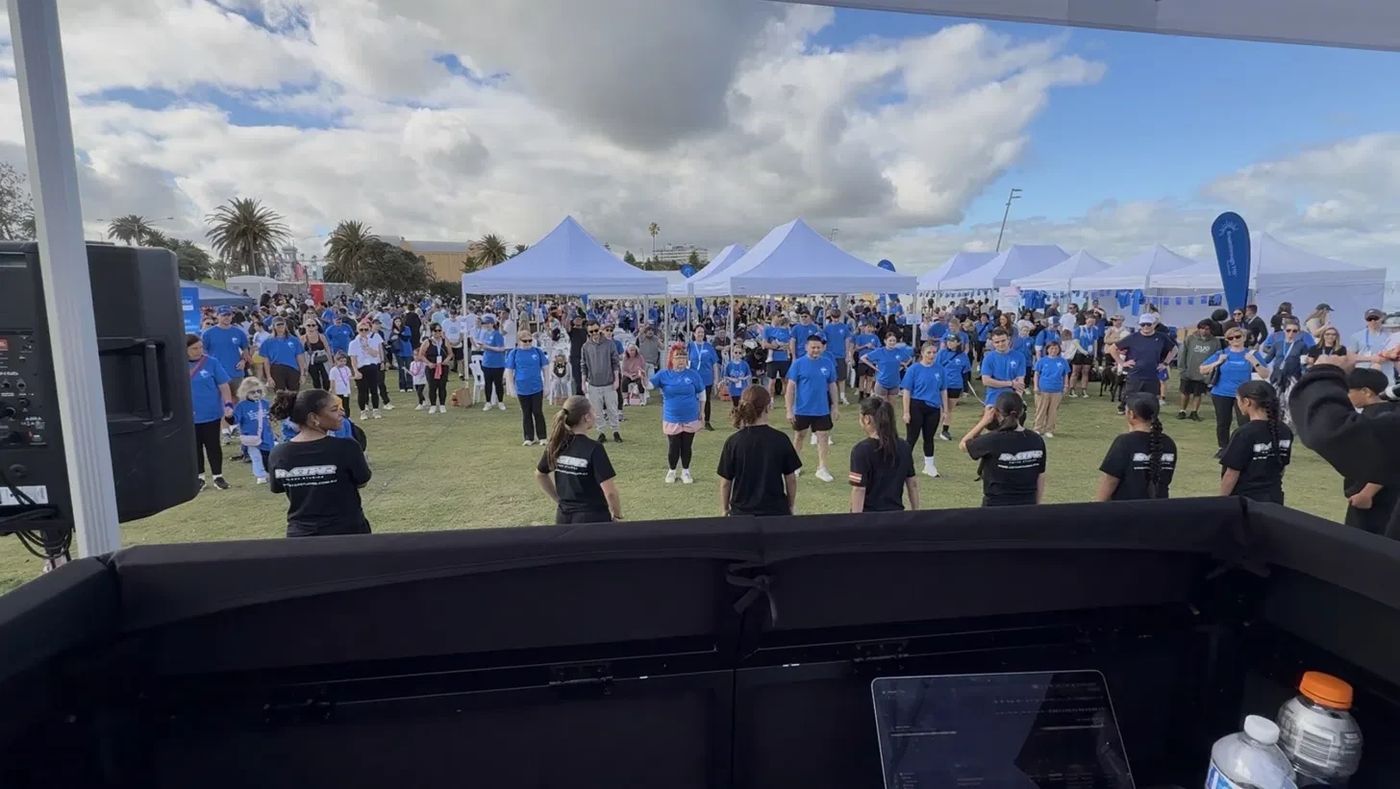 View from the DJ booth at an outdoor community event, with dancers leading a crowd in matching blue shirts